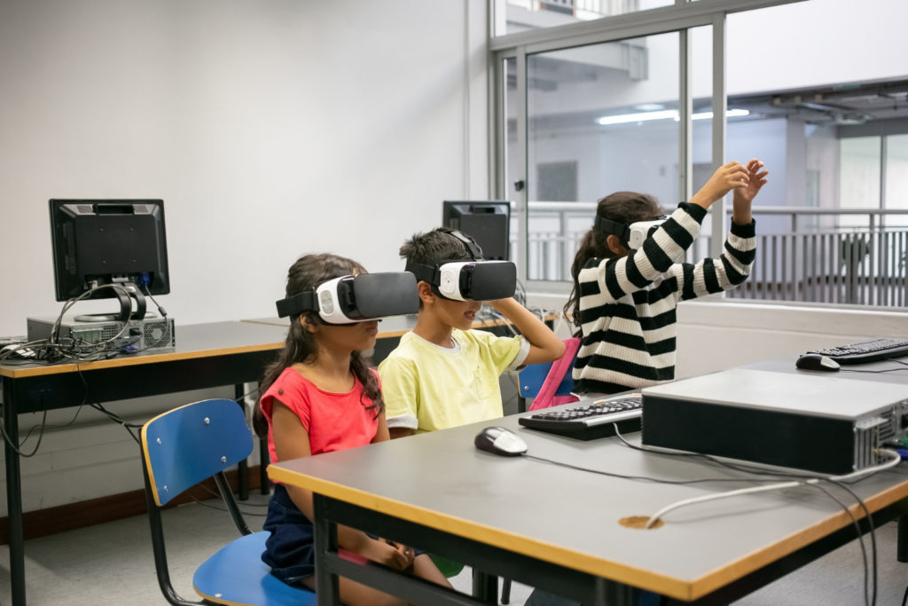La imagen muestra a tres niños en un aula, usando gafas de realidad virtual. Esto indica la integración de la tecnología avanzada en el entorno educativo para mejorar y diversificar los métodos de enseñanza, permitiendo experiencias de aprendizaje inmersivas