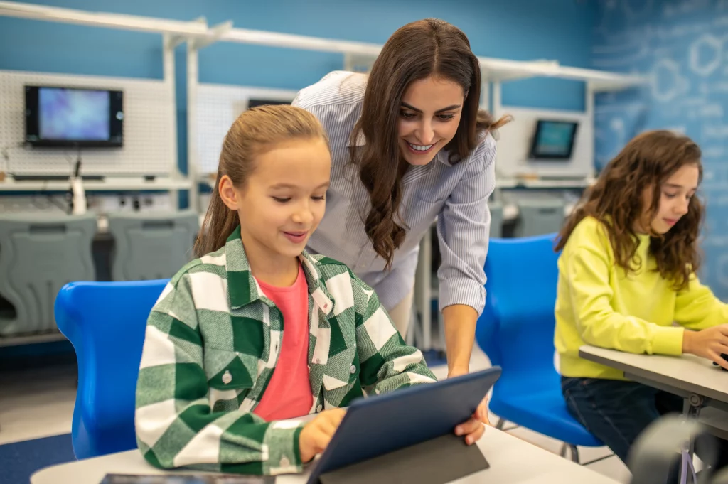 La imagen muestra a un niño en un aula moderna, utilizando una tableta digital mientras recibe asistencia de una persona. La imagen resalta la integración de la tecnología en la educación para facilitar el aprendizaje interactivo y personalizado.
